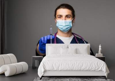 Portrait of medic with hairstyle in lab blue uniform and sterile mask checking, preparing vaccine having syringe in hands, isolated on grey background Wall mural